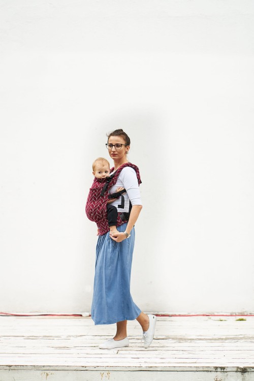 Modelka nosi dziecko w ergonomicznego nosidle KAVKA Multi‑Age burgundy braid cotton - burgundowe nosidło z białym wzorem warkocza