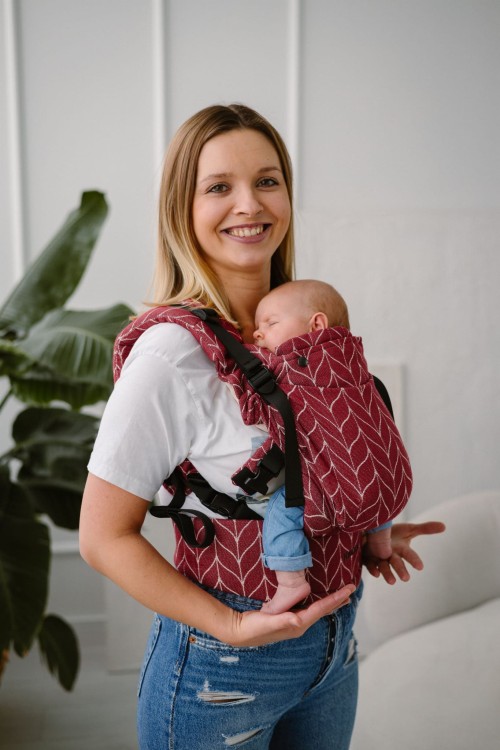 Modelka nosi dziecko w ergonomicznego nosidle KAVKA Multi‑Age burgundy braid cotton - burgundowe nosidło z białym wzorem warkocza