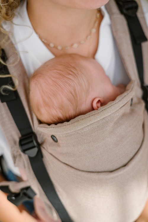 Modelka nosi dziecko w ergonomicznym nosidle KAVKA multi-age walnut twill -  brązowe nosidło z delikatnym wzorem w jodełkę . (ENG: a brown carrier with  twill - subtle herringbone pattern) Przy karku dziecla zamontowany jest tzw. head support