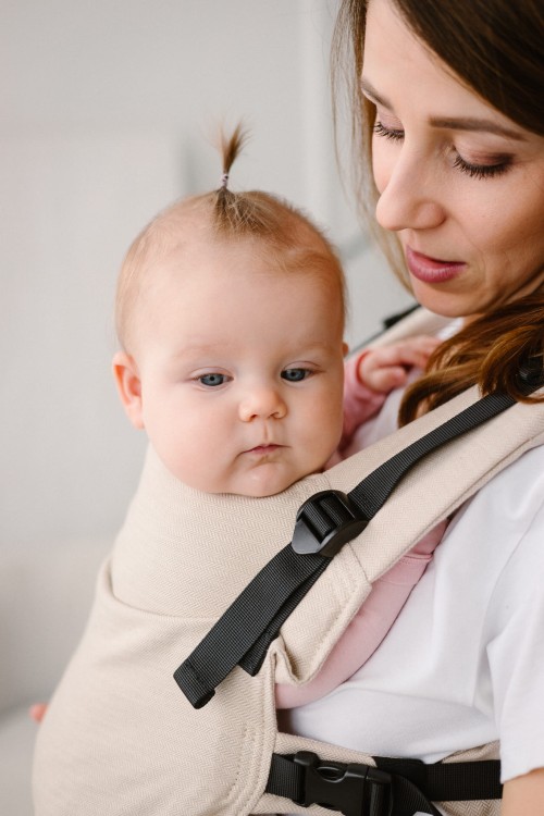 Modelka nosi dziecko w ergonomicznym nosidle KAVKA multi-age vanilla twill  - beżowe nosidło z białym wzorem w drobną jodełkę.   (a beige carrier with a white twill pattern)