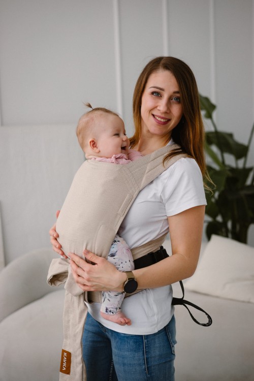 Modelka nosi dziecko w ergonomicznym nosidle KAVKA handy vanilla twill -  beżowe nosidło z białym  wzorem w drobną jodełkę  (a beige baby carrier with white twill  pattern)