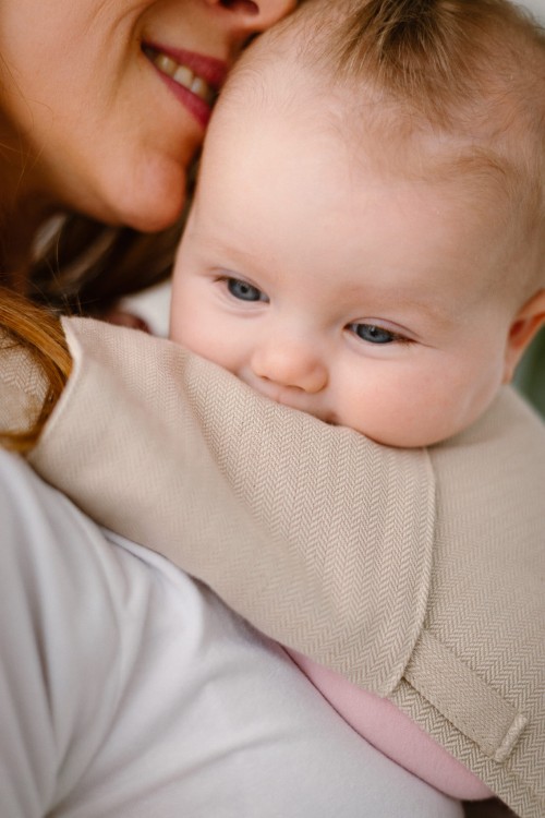 Modelka nosi dziecko w ergonomicznym nosidle KAVKA handy vanilla twill -  beżowe nosidło z białym  wzorem w drobną jodełkę  (a beige baby carrier with white twill  pattern)